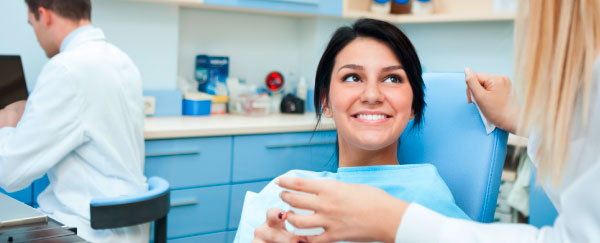 Behandlungssituation: Zahnarzthelferin reicht einer sympathischen Patientin einen Becher Wasser, im Hintergrund ist abgewandt der Zahnarzt zu sehen.